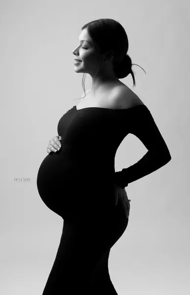 Black and white silhouette of expecting mother in profile in an off-shoulder black gown, eyes closed, hand on her bump, hair in a low bun