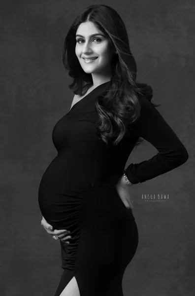 Black and white portrait of smiling expecting mother in a fitted black ruched one-shoulder long-sleeve gown with a high slit, long wavy