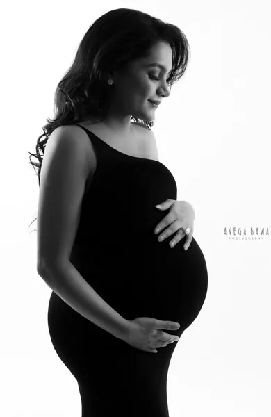 High-key black and white portrait of expecting mother in a black one-shoulder dress, eyes closed, hand on her bump, wavy hair, white