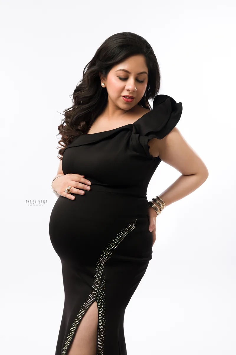 Maternity photoshoot in Delhi: expecting mother in a black one-shoulder gown with a ruffled sleeve and a crystal-trimmed side slit, hand on