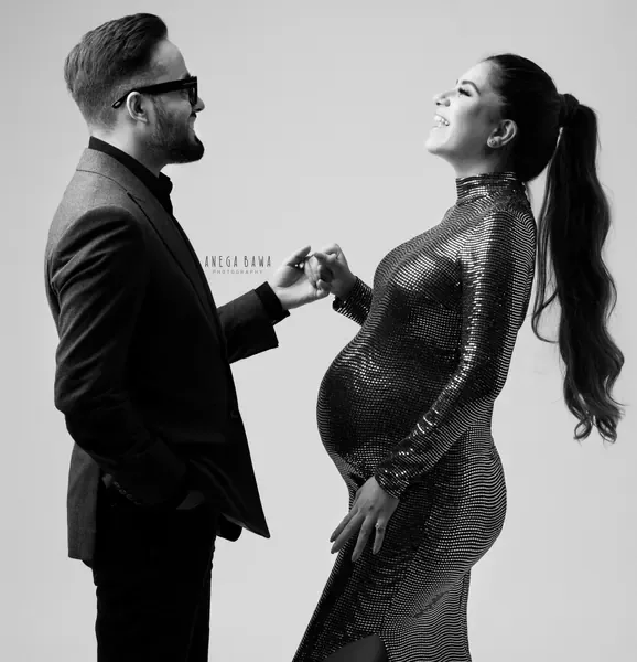 Black and white portrait of a couple, husband in a blazer and glasses holding his laughing wife's hand, she wears a fitted sequin high-neck