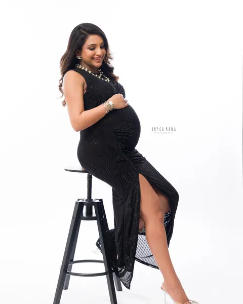 Maternity photoshoot in Delhi: smiling expecting mother seated on a black stool in a sleeveless black studded dress with a side slit and