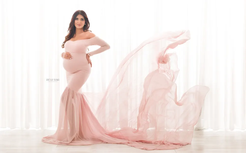 Maternity photoshoot in Delhi: expecting mother in a blush pink off-shoulder mermaid gown with a sheer train blowing out behind her, white
