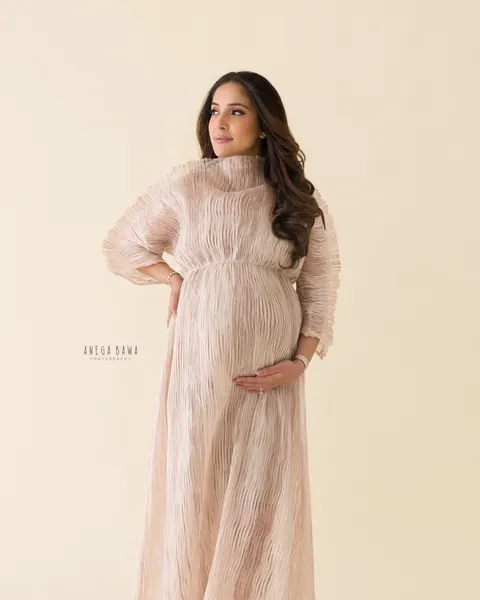Maternity photoshoot in Delhi: expecting mother in a flowing blush pleated high-neck gown with one hand on her hip and one on her bump