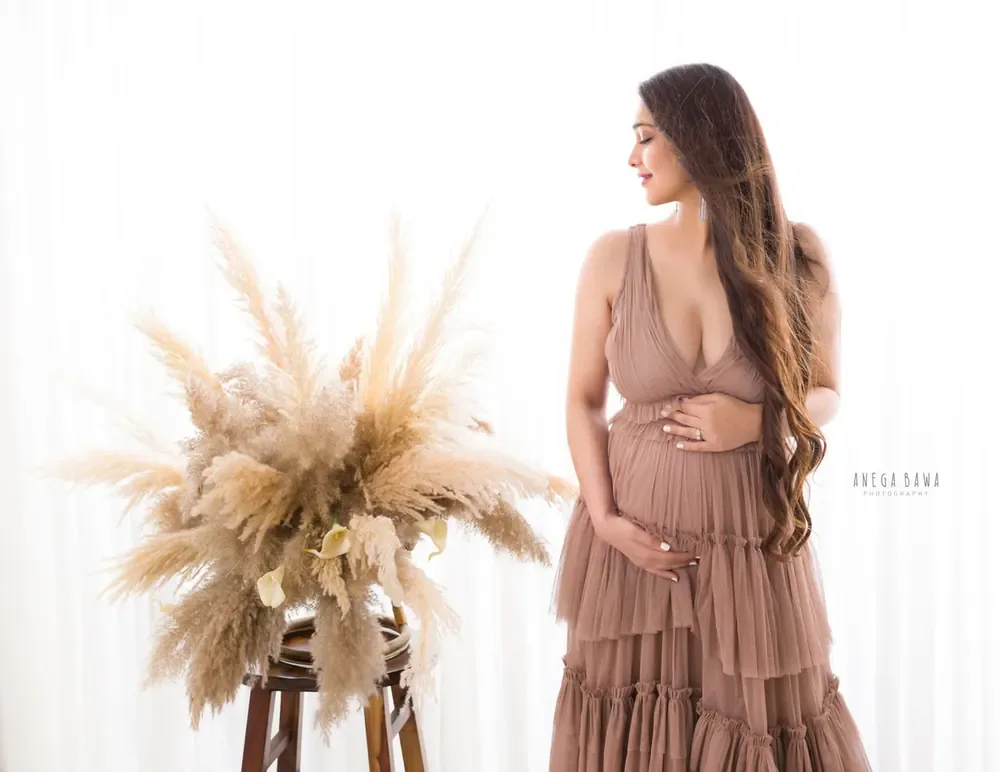 Expecting mother in a mauve tiered ruffle halter gown with a plunging neckline, looking to the side beside a pampas grass arrangement on a