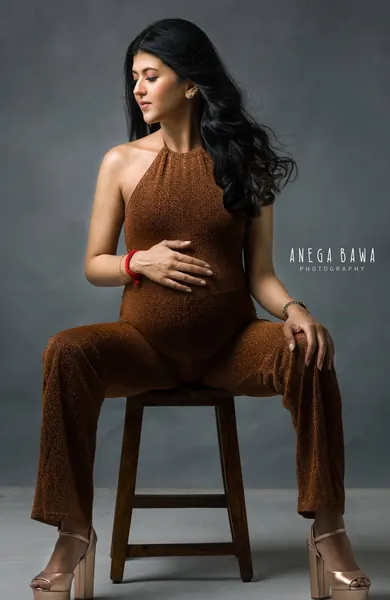 Expecting mother seated on a wooden stool in a rust-brown halter jumpsuit with wide legs and platform heels, hand on her bump, looking to
