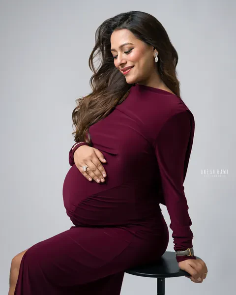 Maternity photoshoot in Delhi: expecting mother in a burgundy long-sleeve gown seated on a black stool with a hand on her bump, grey