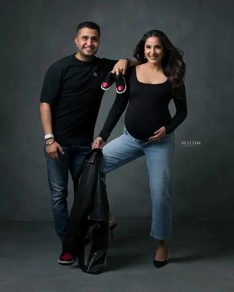 Maternity photoshoot in Delhi: smiling couple in black tops and jeans, husband holding a leather jacket and a pair of tiny red and black