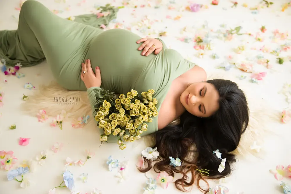 Overhead view of expecting mother lying on a fur rug in a sage green long-sleeve gown, holding a bouquet of yellow roses, surrounded by