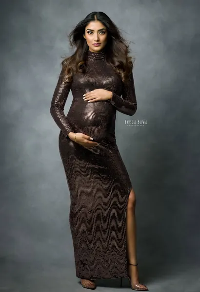 Expecting mother in a fitted bronze sequin high-neck long-sleeve gown with a thigh slit and clear heels, hands on her bump, grey-blue