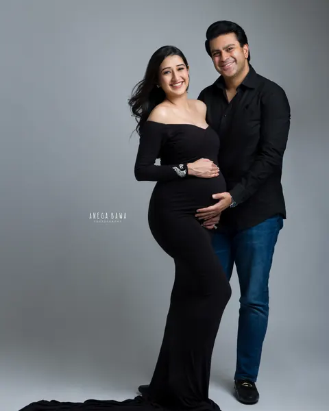 Maternity photoshoot in Delhi: smiling couple, wife in a fitted off-shoulder black gown with a long train and husband in a black shirt and