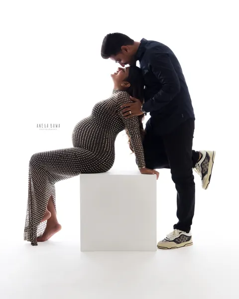 Maternity photoshoot in Delhi: husband in a navy shirt leaning down to touch foreheads with his wife who sits on a white block in a studded