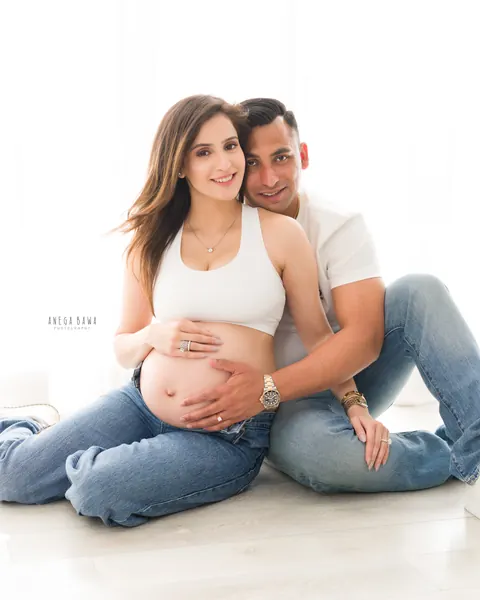 Maternity photoshoot in Delhi: smiling couple seated on a white floor in white tops and denim, husband's hand on her bare bump, by Anega