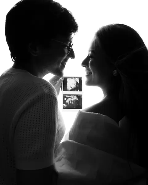 Maternity photoshoot in Delhi: black and white silhouette of a couple smiling at each other while holding ultrasound scan photos, by Anega