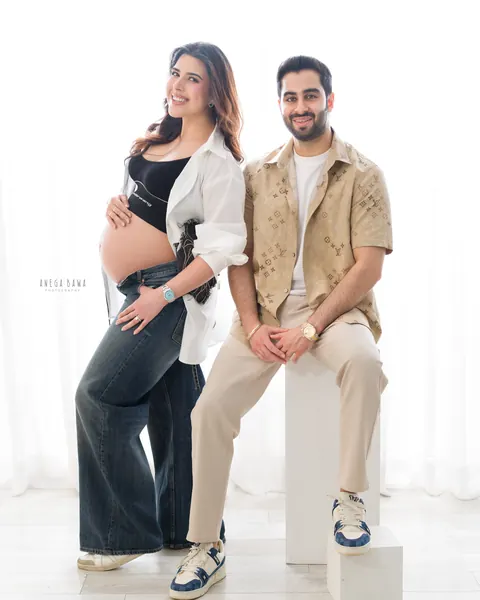 Maternity photoshoot in Delhi: smiling couple, husband seated on a white block in a patterned shirt and beige trousers and wife standing in