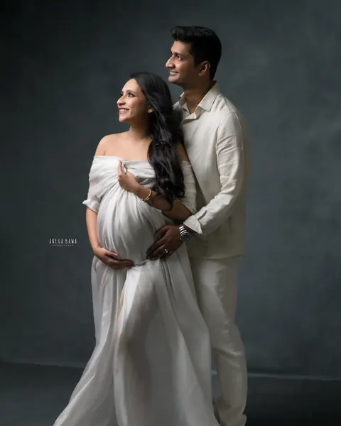 Maternity photoshoot in Delhi: couple in white looking up together, wife in a flowing sheer white drape with a long train and husband in a