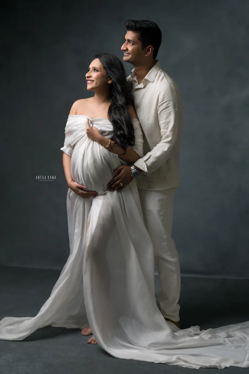 Maternity photoshoot in Delhi: couple in white looking up together, wife in a flowing sheer white drape with a long train and husband in a