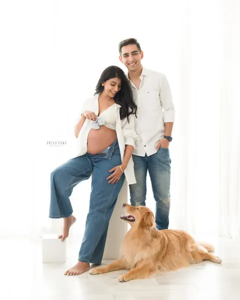 Maternity photoshoot in Delhi: smiling couple in white shirts and jeans with their golden retriever lying at their feet, wife holding baby