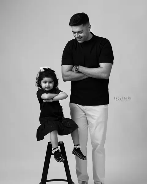 Maternity photoshoot in Delhi: black and white portrait of a father and his toddler daughter both with arms crossed, she sits on a black