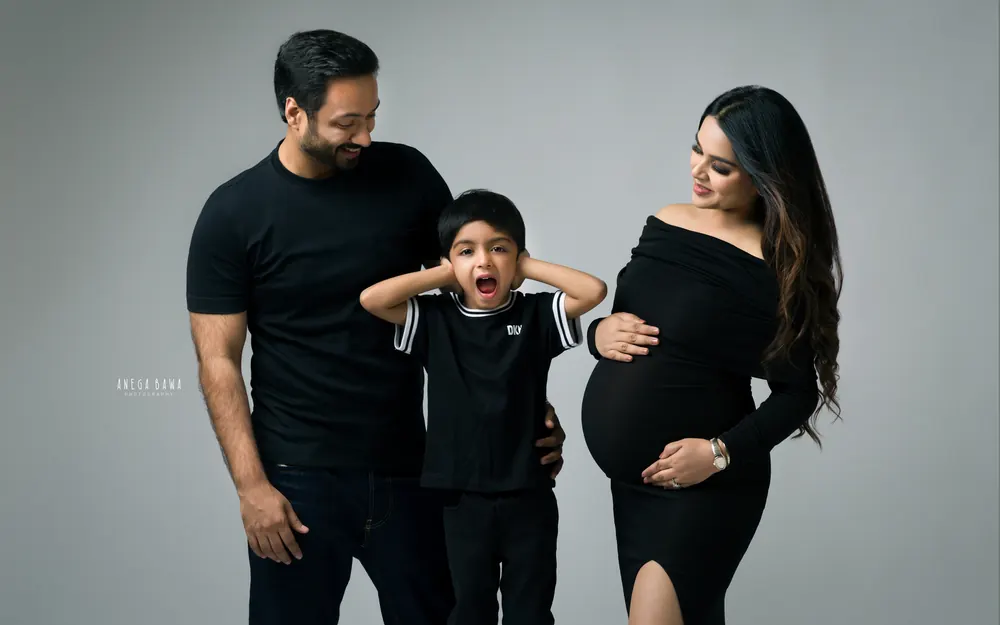 Maternity photoshoot in Delhi: playful family in black, young son covering his ears and yawning between his father and expecting mother