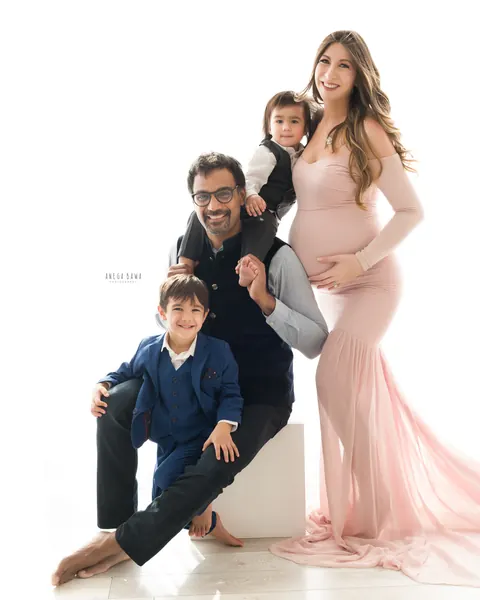 Maternity photoshoot in Delhi: family with two sons, mother in a blush pink gown with a sheer train, father seated on a white block with