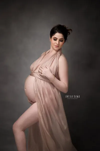 Expecting mother in a blush chiffon halter drape with a high slit, hair in a messy updo, hand on her chest, dark moody grey backdrop.
