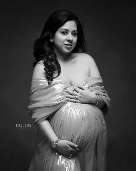 Maternity photoshoot in Delhi: black and white portrait of expecting mother wrapped in a metallic drape, both hands cradling her bump