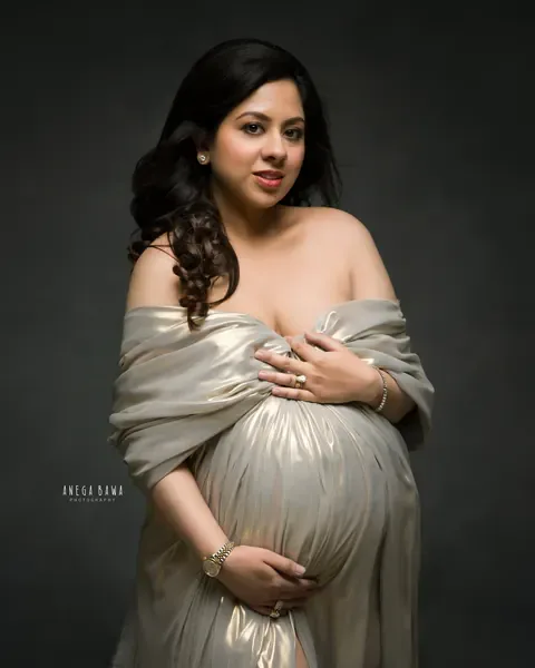 Maternity photoshoot in Delhi: expecting mother wrapped in a gold metallic drape off the shoulders, both hands cradling her bump, looking