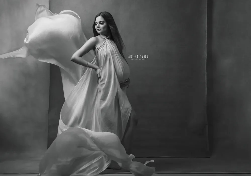 Black and white portrait of expecting mother in a draped halter satin gown with sheer fabric flying beside her, hand on hip, textured grey