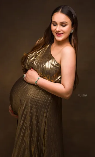 Smiling expecting mother in a gold metallic pleated one-shoulder gown with drop earrings, looking down, hands cradling her bump, warm brown