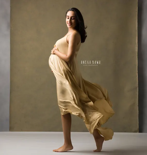 Smiling expecting mother in a champagne satin slip dress with the skirt flowing as she steps, barefoot, hand on her bump, olive and grey