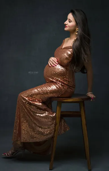 Expecting mother seated on a wooden stool in a copper sequin fitted gown with a fishtail hem and heels, looking away with a hand on her