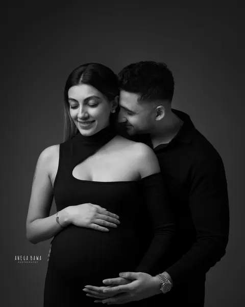 Maternity photoshoot in Delhi: black and white portrait of a husband nuzzling his smiling wife's neck from behind, she wears a black knit