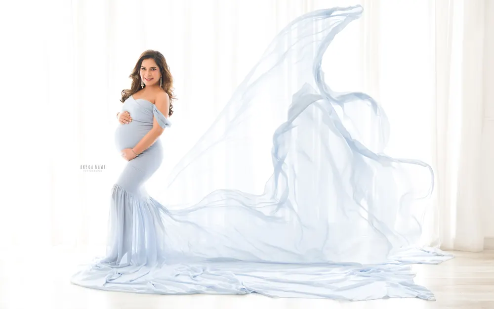 Maternity photoshoot in Delhi: expecting mother in a light blue off-shoulder mermaid gown with a huge sheer train billowing behind her
