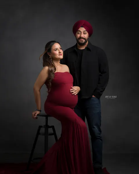 Maternity photoshoot in Delhi: expecting mother seated on a stool in a maroon mermaid gown with a long train, husband standing behind in a