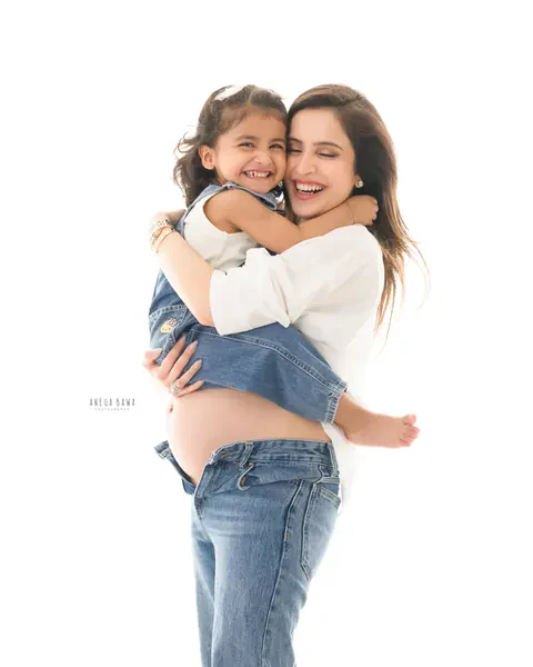 Maternity photoshoot in Delhi: laughing mother in a white shirt and denim carrying her toddler daughter in dungarees, bare bump showing