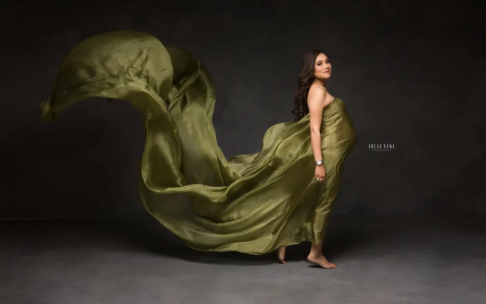 Maternity photoshoot in Delhi: expecting mother walking barefoot in an olive green drape with a long trail of fabric behind her, dark