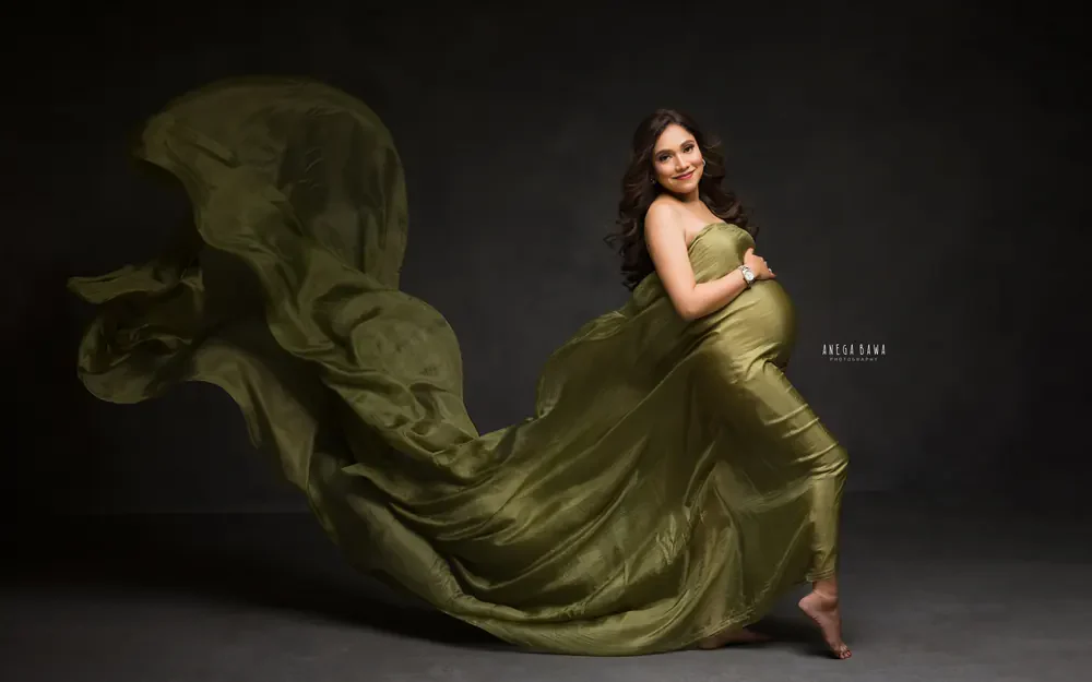 Maternity photoshoot in Delhi: expecting mother in an olive green satin drape flowing behind her against a dark studio backdrop, by Anega