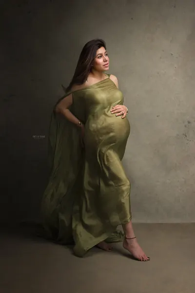 Expecting mother in an olive green sheer drape pinned at one shoulder, barefoot, hand on hip and one on her bump, looking up, textured