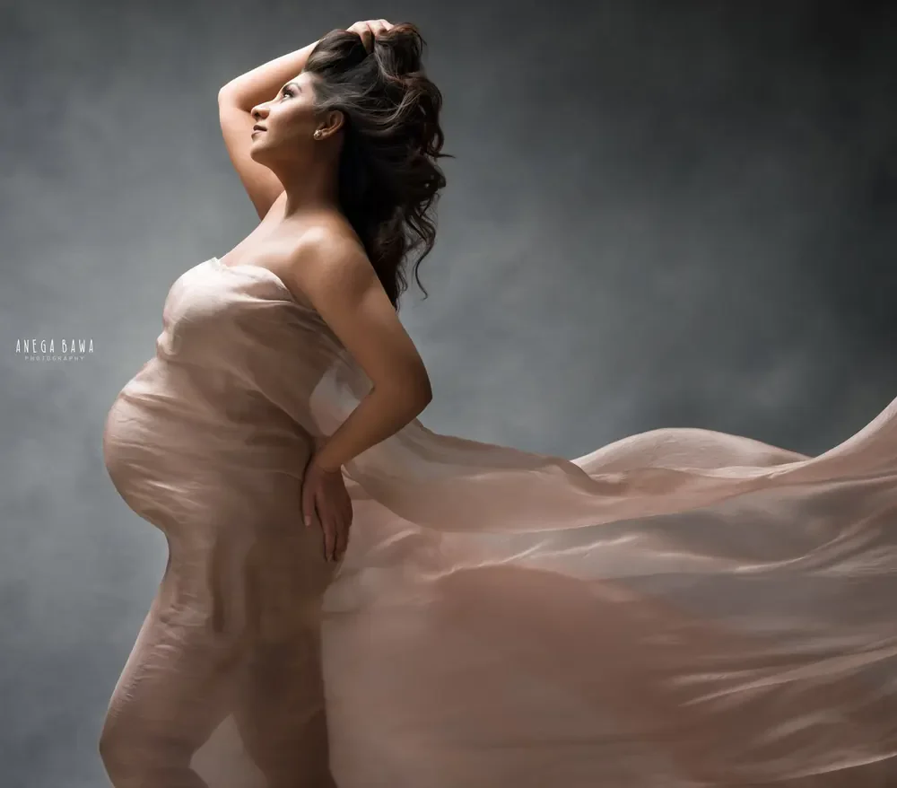 Expecting mother in a blush silk drape across her chest with fabric streaming out behind her, one hand in her hair and one on her hip