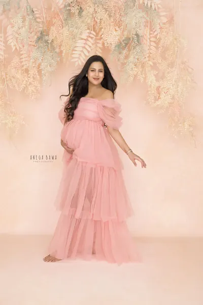 Expecting mother in a pink tiered tulle off-shoulder gown with puffed sleeves, barefoot, hand on her bump, standing under hanging peach