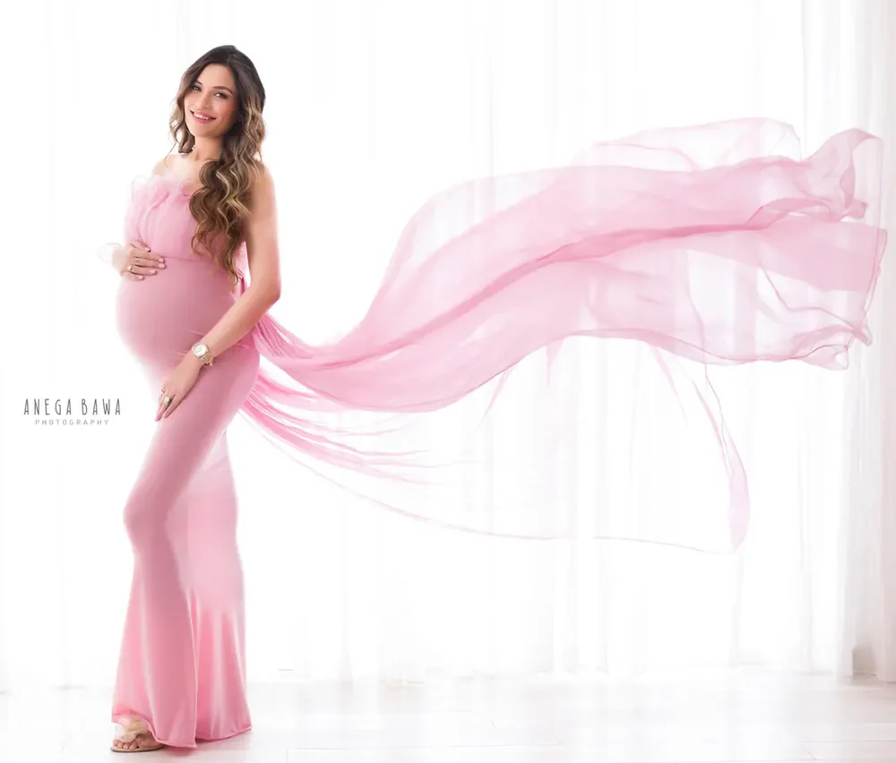 Expecting mother in a rose pink strapless fitted gown with a sheer pink scarf streaming out behind her, hand on her bump, bright white
