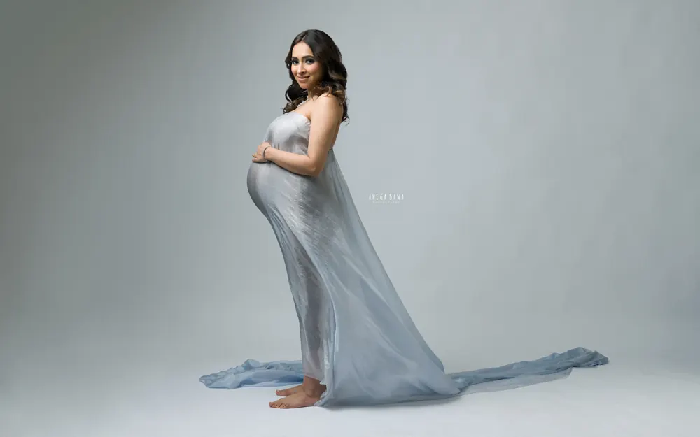 Maternity photoshoot in Delhi: expecting mother smiling in a strapless powder blue silk drape with a long train on the floor, soft grey