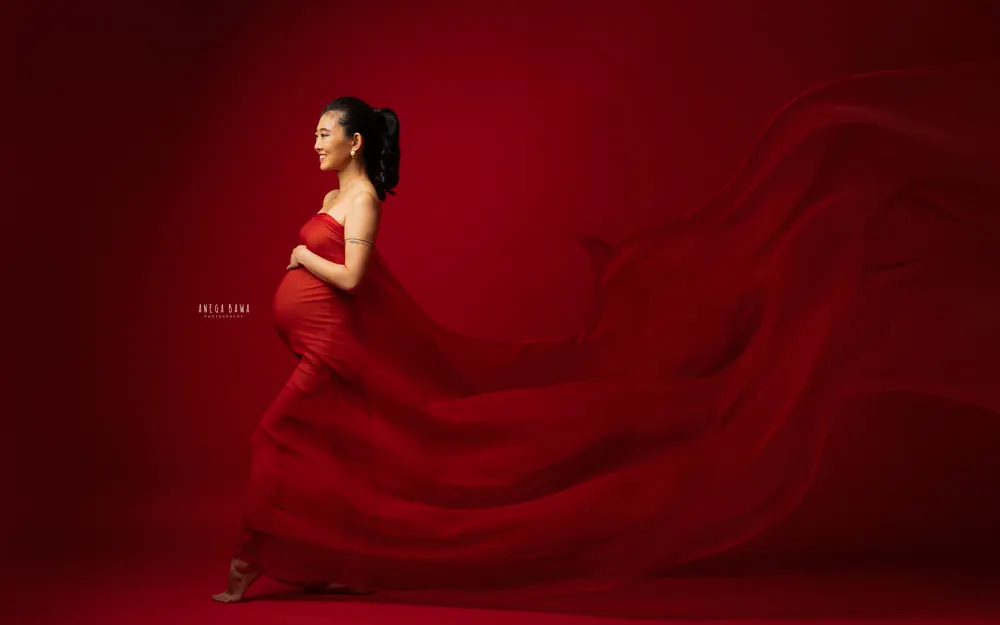 Maternity photoshoot in Delhi: expecting mother in profile walking barefoot in a strapless red drape with a long flowing train, deep red