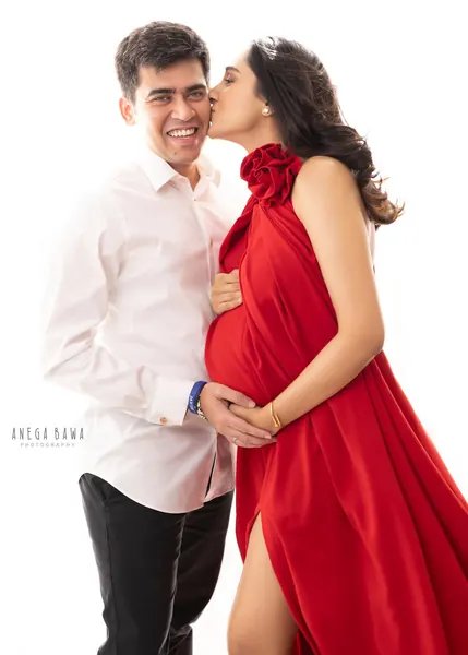 Smiling couple, husband in a white shirt holding his wife's bump while she kisses his cheek, wife in a red halter gown with a rose at the