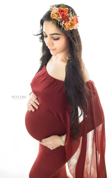 Expecting mother in a maroon fitted gown with a sheer cape sleeve and an orange and red flower crown, long dark hair, looking down, hands