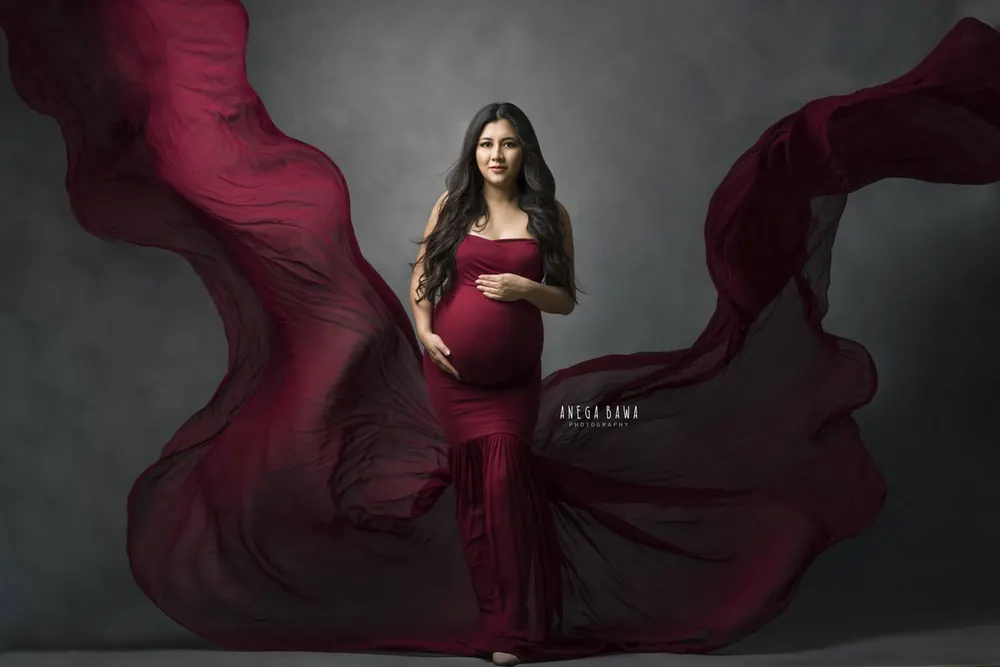 Expecting mother in a burgundy strapless mermaid-style gown with sheer burgundy fabric billowing out on both sides, one hand on her bump