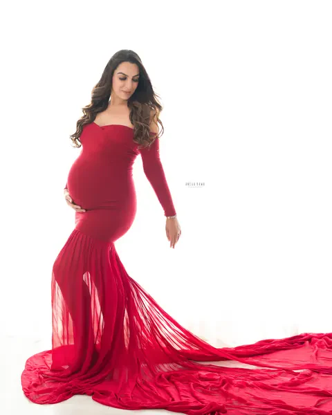 Maternity photoshoot in Delhi: expecting mother in a red off-shoulder mermaid gown with a sheer flowing train across a bright white studio