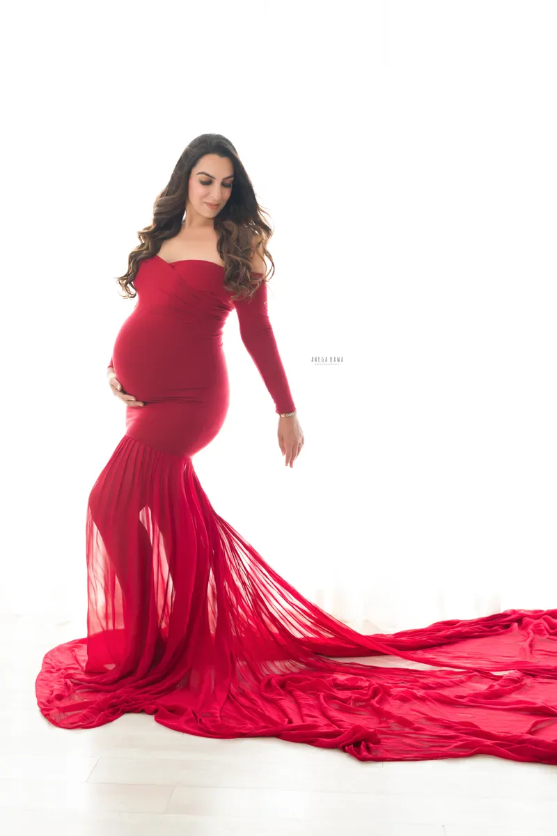 Maternity photoshoot in Delhi: expecting mother in a red off-shoulder mermaid gown with a sheer flowing train across a bright white studio