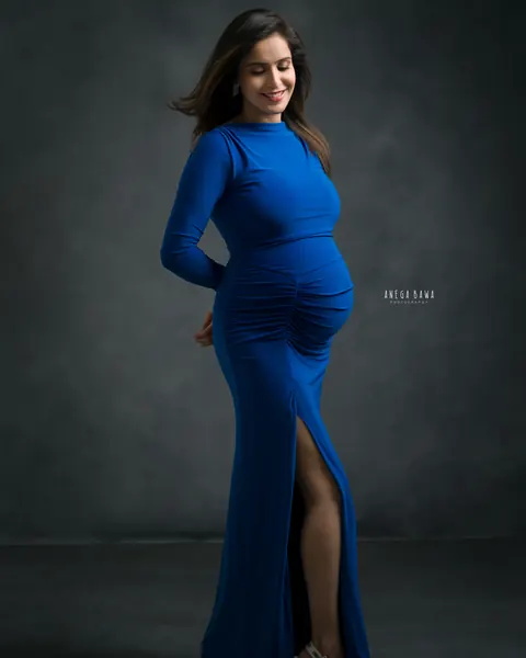 Maternity photoshoot in Delhi: expecting mother in a royal blue ruched long-sleeve gown with a side slit and nude heels, hands behind her